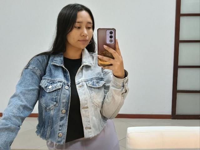 Karollteyes with long black hair, wearing a light blue denim jacket and black shirt, holding a phone in her right hand, taking a mirror selfie in a bathroom.