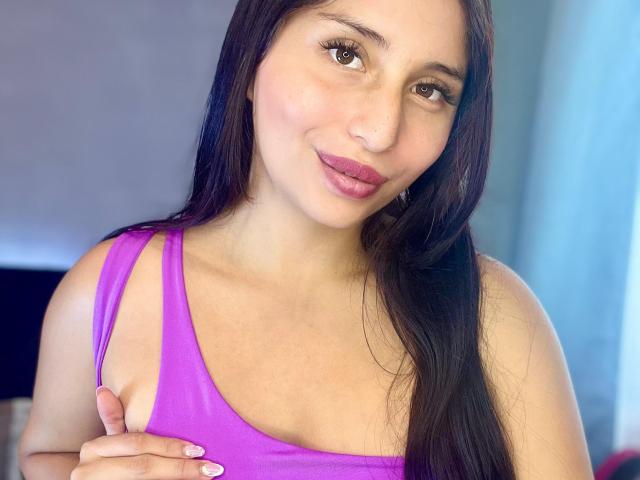 KateOwens has long black hair, brown eyes, and pink lipstick, wearing a purple tank top, holding her left tit with her right hand, smiling softly.