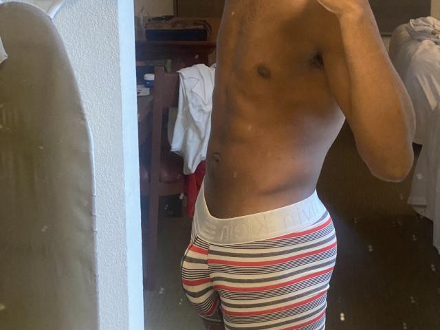 Shirtless KingBlaze with a toned, brown-skinned, muscular, flat-chested body, wearing striped, red, white, and black boxer briefs, standing in front of a mirror, taking a selfie.
