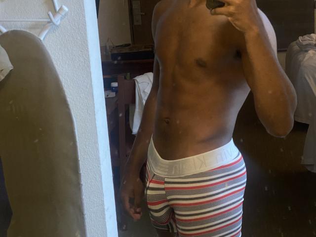 O torso musculoso e sem camisa de KingBlaze, com pele escura e usando cuecas boxer listradas em vermelho, branco e cinza, exibe um bumbum visível, proeminente e bem formado. Sua mão segura um telefone, tirando uma selfie no espelho..