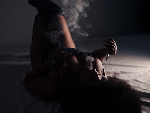 AliceJelanni is lying on a bed, smoking, with a wisp of smoke around her face. She's got dark skin, afro hair, and is wearing black lingerie.