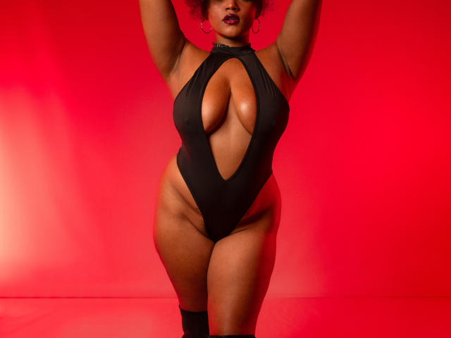 AliceJelanni in a black cutout one-piece, showing her big boobs and thick thighs, with arms raised, wearing red lipstick and hoop earrings, against a red background.