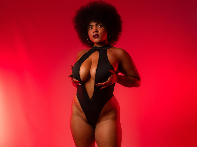 AliceJelanni with big afro, wearing a black cutout one-piece, holding her big tits, standing against a red background, looking confident. Her dark skin contrasts with the red.
