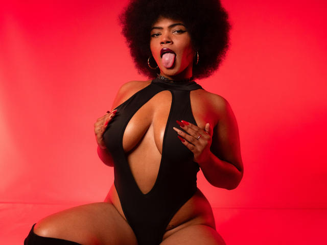 AliceJelanni with big afro, sticking out tongue, wearing black cutout one-piece, hands on huge tits, red background, red nails, hoop earrings, sitting with legs apart.