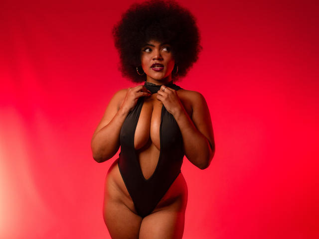 AliceJelanni with big afro, wearing a black deep-cut one-piece, holding the collar, showing her big boobs and thick thighs, against a red background.