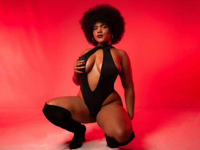 Alice Jelanni, com um afro grande, vestindo um macacão preto com recortes, ajoelhada, com as mãos nos seios, botas de cano alto, fundo vermelho, expressão confiante, batom vermelho escuro, brincos de argola..