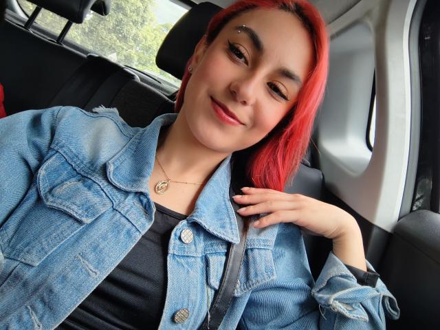 AmyStreetwise69 with red hair, wearing a denim jacket and black top, smiling in a car, hand on chest, small nose piercing, gold necklace, cute, sexy, casual.