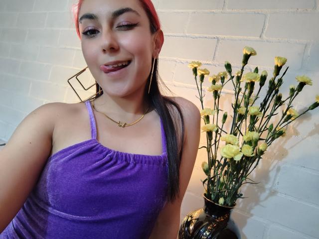 AmyStreetwise69 with pink hair, purple dress, and gold necklace, smiling with slightly parted lips, showing her tits, holding a vase of yellow flowers on a white brick wall.