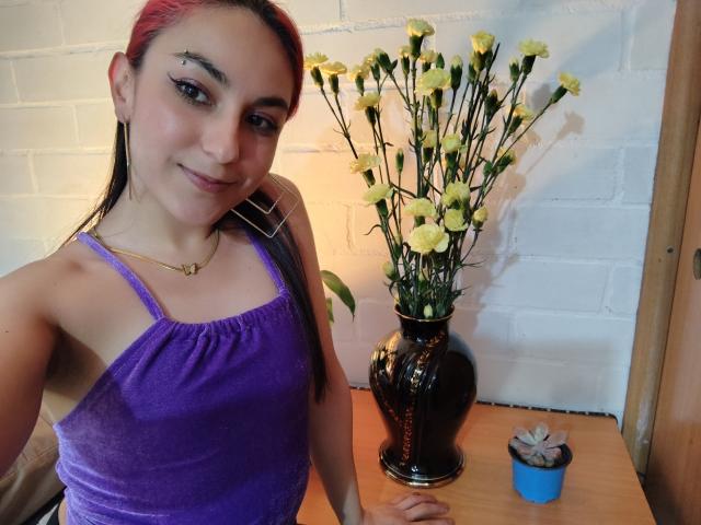 AmyStreetwise69 with pink-dyed hair, purple velvet top, small breasts, black vase with yellow flowers, gold necklace, and a blue plant pot, smiling at the camera.