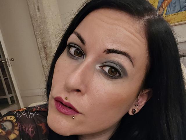 GennyRock has dark hair, gray eye makeup, a nose piercing, and a lower lip piercing. She's wearing purple lipstick and black ear studs, looking slightly up with a subtle smile.