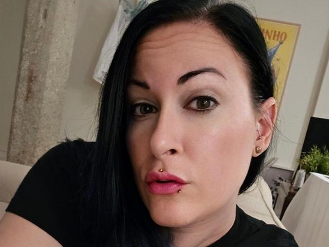 GennyRock has black hair, brown eyes, and a black shirt. She has a nose ring, a lower lip piercing, and pink lipstick. Her face is slightly squished.