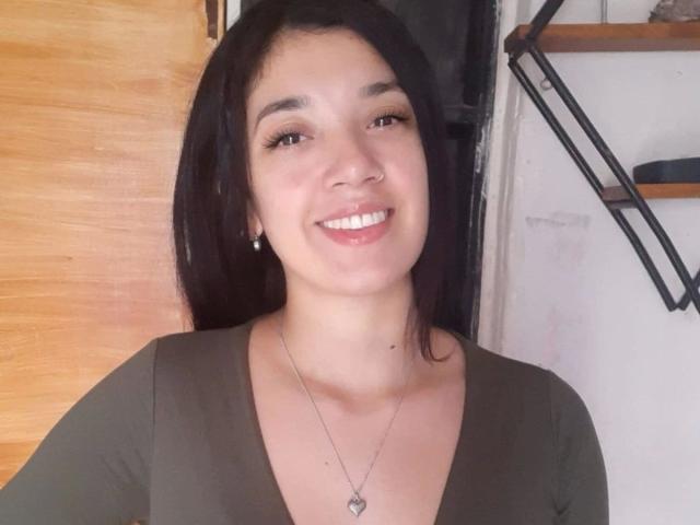 CamySensual-hot with dark hair, brown eyes, and a smile, wearing a green V-neck top, necklace with heart pendant, and earrings, standing in front of a wooden door.