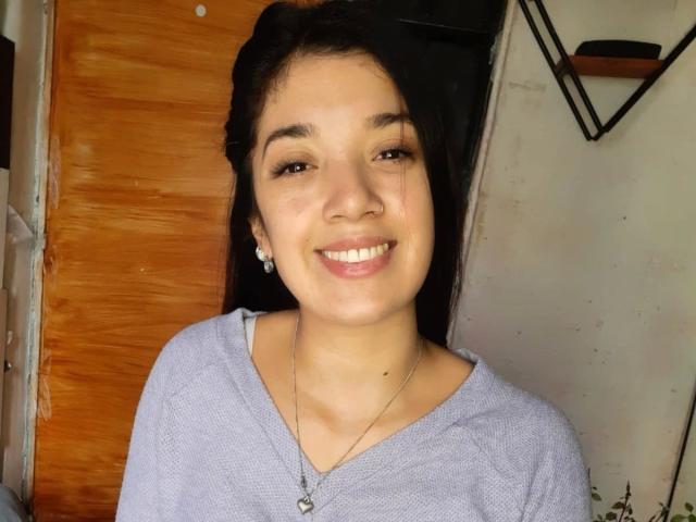 CamySensual-hot with dark hair, light skin, brown eyes, smiling, wearing a gray shirt, necklace, earrings, standing against a wooden door, and a white wall in the background.