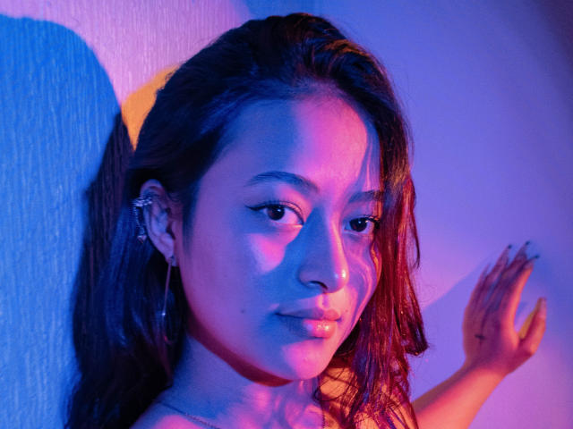 SamaraCuttie has dark brown hair, medium skin, and is wearing a nose ring with hoop earrings. She's in blue and pink lighting, with her hand against a wall.