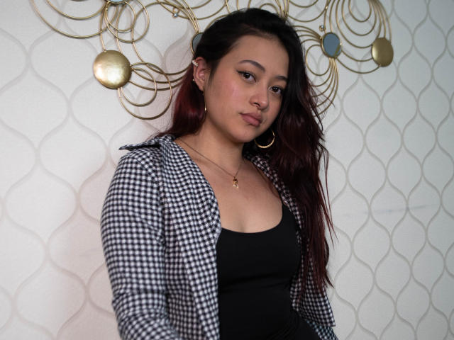 SamaraCuttie with dark red hair, wearing a black and white checkered jacket, black tank top, and hoop earrings, has a small gold necklace and a subtle nose ring.