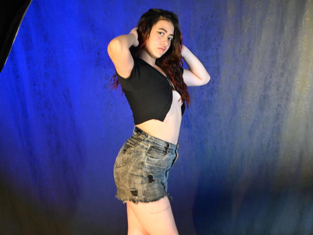 SamaraCuttie with long red hair, black crop top exposing her tits, and frayed denim shorts, standing sideways, hands in hair, blue and gray background, looking at camera.