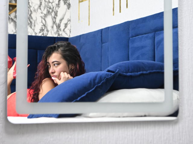 SamaraCuttie with red hair, lying on a blue-pillow bed, resting her face on her hand, wearing a red top, looking sideways, seen through a white-framed window.