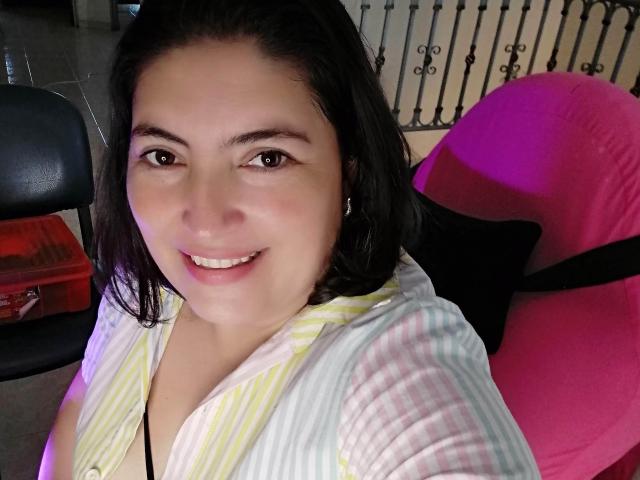 Lina69s with dark hair, smiling, wearing a striped yellow and white shirt, sitting in a pink chair, black strap visible, black hair clip, pink and purple light.