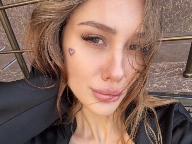 SensualMila with pouty lips, light brown hair, and a small heart tattoo under her right eye. She's wearing a black jacket, with long eyelashes and a slight smile.