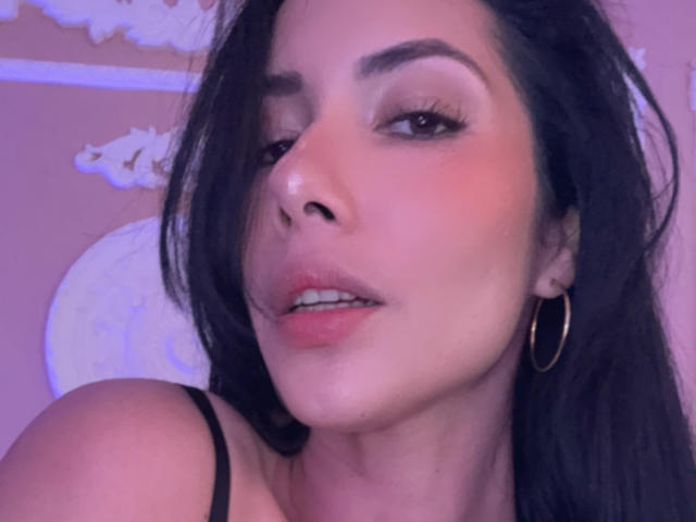 ArianaRogers with pouty pink lips, dark eyebrows, long black hair, gold hoop earrings, and a slight nose piercing, wearing a black strap, in a purple-lit selfie.
