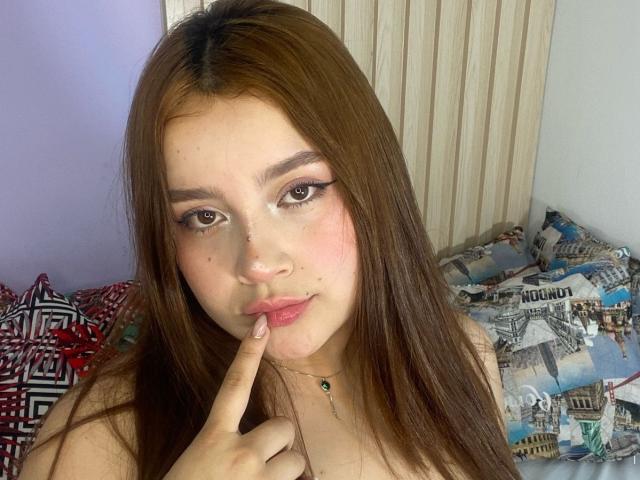 ZairaVanCleef mit langen braunen Haaren, rosafarbenen Lippen und einem Finger in der Nähe ihres Mundes, die ihr süßes, mit Sommersprossen übersätes Gesicht und ihr dezentes Make-up zur Geltung bringt. Sie trägt eine dünne, grüne Halskette..