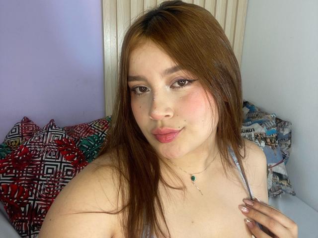 ZairaVanCleef, com cabelo castanho, lábios rosados e olhos verdes, usando um colar, segurando uma alça, mostrando os ombros nus e com uma pequena piercing no nariz, sorrindo para a câmera..