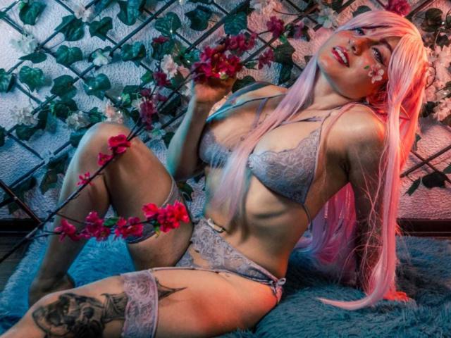 MeiJaisen with pink hair, lace lingerie, and tattoos, holding red flowers, sitting on a blue furry rug, legs spread, showing her tits and pussy, in a sexy pose.