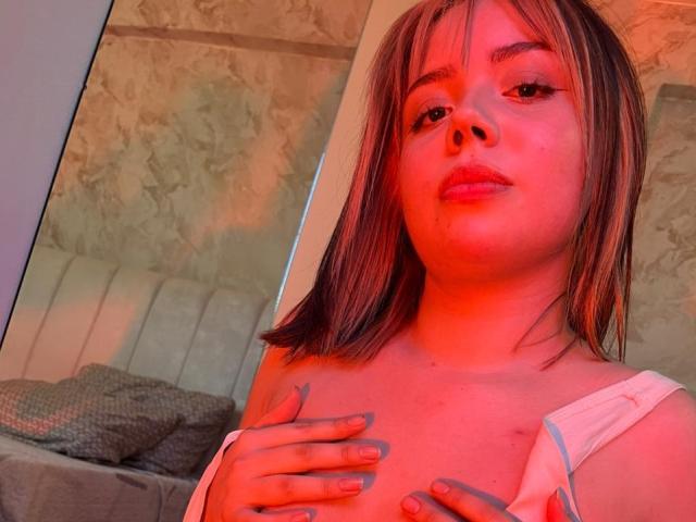 HanniRoss with red-tinted skin, medium brown hair, and pink lipstick, covering her tits with hands, wearing a white top, in a dimly lit room.