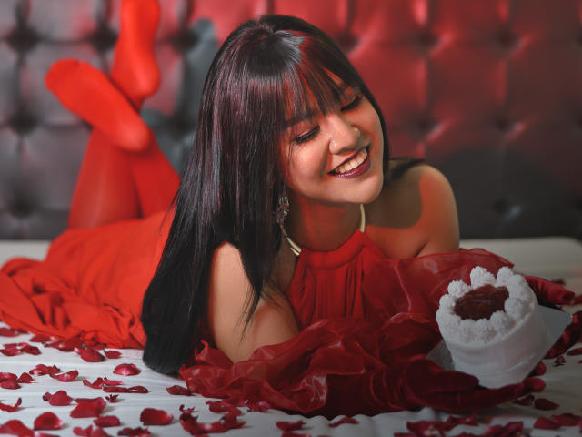 AlluringAmaraTS, with long black hair and red lipstick, lies on a bed, smiling, wearing red stockings and a red dress, holding a heart-shaped cake with red rose petals.