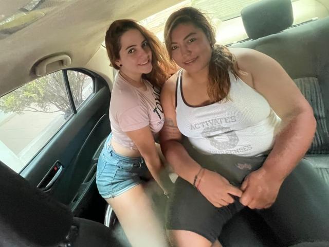 Estefany with small tits and short denim shorts, sitting next to Camila with large tits and a white tank top, in a car. Estefany's arm is around Camila's shoulder.