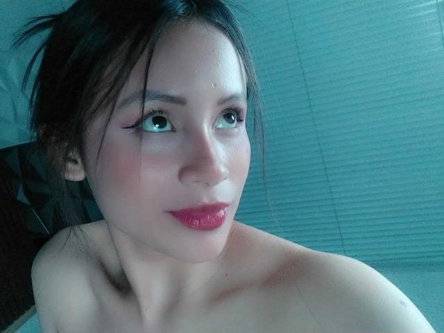TinyLaurent with wet face, dark hair, and red lipstick, looking up with big eyes, bare shoulders visible, blue-green light. TinyLaurent's face has a slight blush and she's cute.