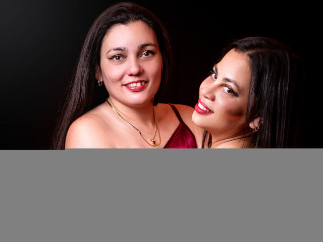 SaharaMK is kissing SaharaMK's neck while SaharaMK lies on her back, wearing a red satin bra, with her tits and dark hair visible, in a dark, intimate setting.