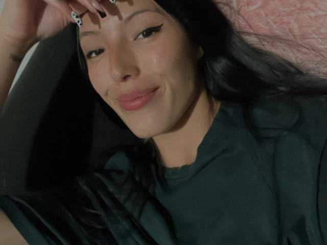 Luluberrie in a sheer black lingerie with red trim, showing her ass and tits, sitting on a bed with white pillows and pink blanket. Her hand touches her face.