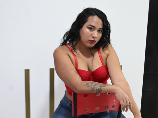 MillenaCollins with wavy black hair, red bustier, showing her tits, tattoo on right arm, and blue jeans unbuttoned, standing in front of a mirror.
