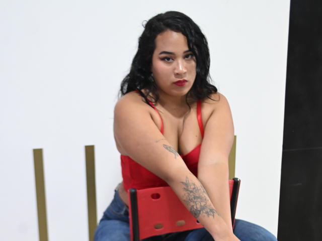 MillenaCollins with black curly hair, red bra, and blue jeans, sitting on a red chair, tattoo on right arm, looking at camera, red lipstick, medium tits.