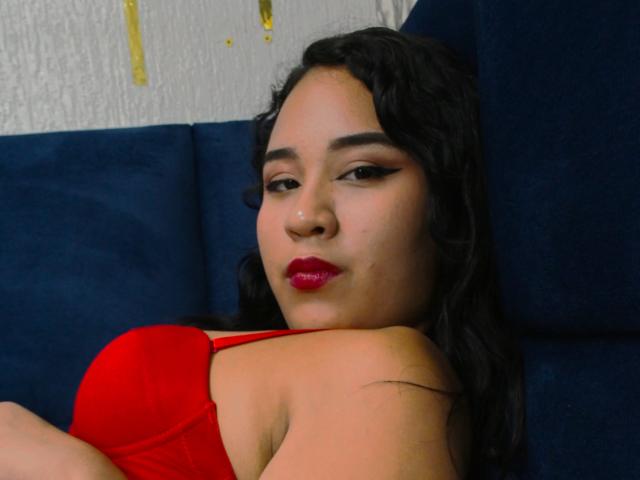 MillenaCollins is on a bed, kneeling with her back to the camera, wearing a red bra, blue jeans, and red high heels, showing her ass. She's looking over her shoulder.
