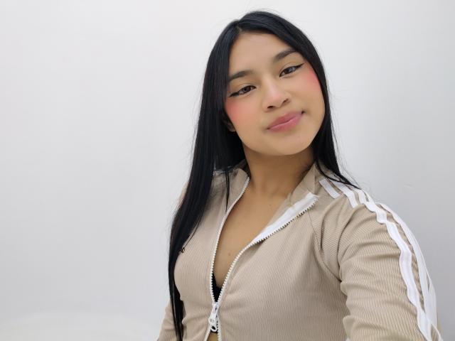 Xihomara with long black hair, wearing a fuzzy beige jacket, exposing black bra, cute pink blush, smiling, showing her tits, against a white background.