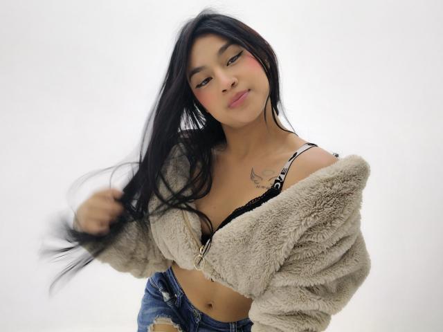 Xihomara with long black hair, pouty lips, tan top, and blue jeans, showing her ass, standing against a white background. Her hands are behind her back.
