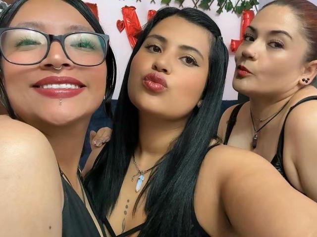 Three SweetFourWomen with black hair, glasses, and red lipstick, pouting, wearing black tops, showing their tits, and a nose piercing, in a selfie with green eye makeup.