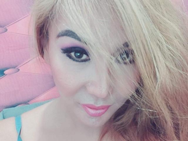 QueenFhukelya, con cabello rubio, delineador de ojos negro intenso, sombra de ojos rosa y labial rosa, tiene un pequeño piercing en la nariz y está sonriendo, con una pequeña gota de líquido blanco en el mentón..