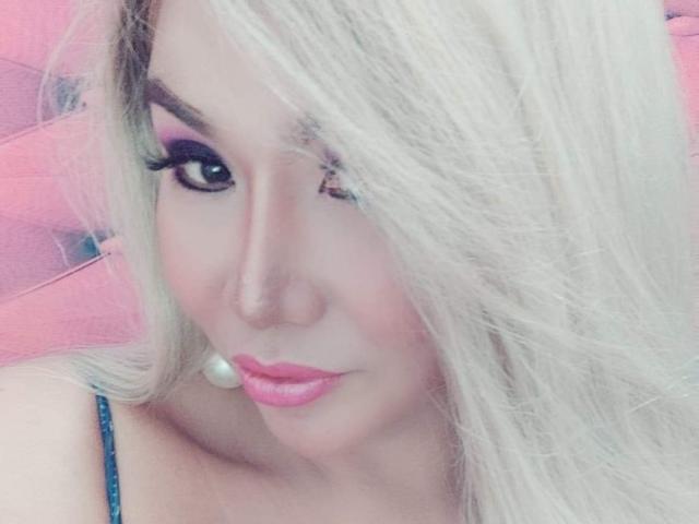 QueenFhukelya, con el cabello rubio cubriendo un ojo, lápiz labial rosa y delineador de ojos oscuro, lleva aretes de perlas y una correa negra, y tiene un aspecto lindo y sexy..