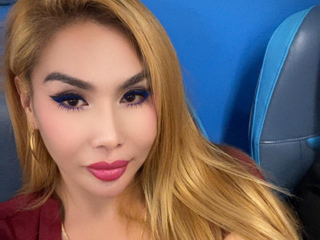 QueenFhukelya hat blondes Haar, blaue Eyeliner, pinken Lippenstift und trägt ein rotes Hemd, mit einem blauen und grauen Hintergrund. Ihre Brüste sind nicht sichtbar..