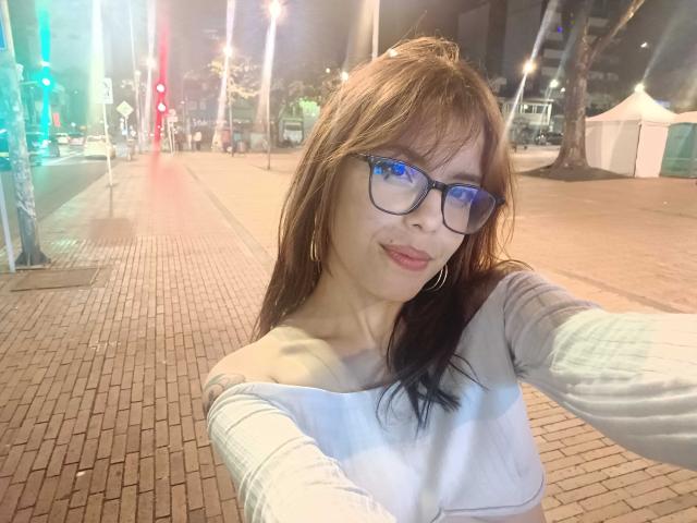 LexyMariee with blue-framed glasses, brown hair, and hoop earrings, wearing an off-shoulder white top, taking a selfie at night on a brick-paved street.
