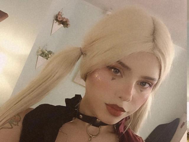 LexyMariee with blonde pigtails, red lipstick, shiny face, black choker, triangle tattoos, and a tattoo on her arm, looking at the camera. Her boobs are partially visible.