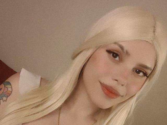 LexyMariee has blonde hair, eyeliner, and a tattoo on her left shoulder. She's smiling with red lips, and her ass is slightly visible. Soft, warm lighting.