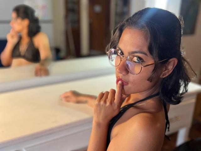 SashaSexyG with dark hair, glasses, and a black halter top, finger to lips, looking in mirror, reflection shows her back, she has a tattoo on her right arm.