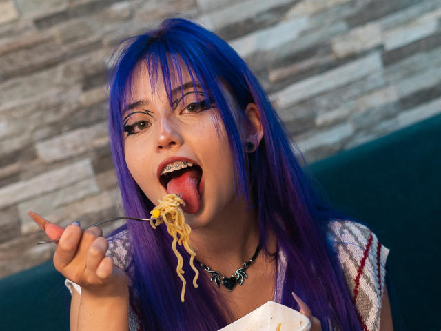 AmeliaXWalker with purple hair, tongue out, holding a fork of noodles, wearing a black choker, earrings, and white shirt, looking at the camera. Her face is slightly messy.