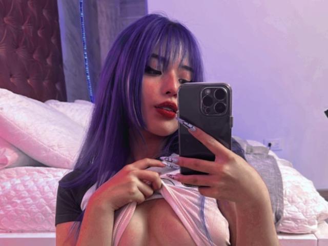 AmeliaXWalker with purple hair, red lipstick, and black eyeliner, lifting her shirt to expose her big tits in a mirror selfie, holding a smartphone with three cameras.