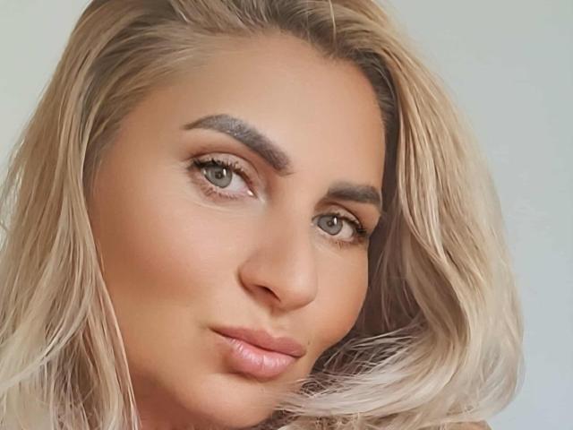 ChaudeEvely has blonde hair, green eyes, and full pink lips. She's looking at the camera with a slight smile, and her eyebrows are dark and well-groomed.