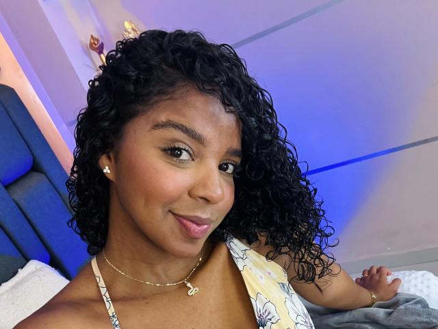 LisaMendez has curly black hair, brown skin, and wears a floral top, showing her small tits, with a gold necklace and earrings, smiling slightly with pink lips.